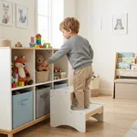 Toddler standing on wooden 2 step stool to reach toys on storage shelf