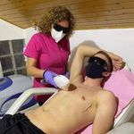A healthcare professional in pink attire and protective mask using an IPL beauty machine on a male client lying down wearing dark eye protection