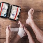 Person using items from an open portable medical kit to bandage an injured foot on a wooden floor